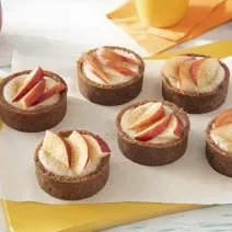 Foto da receita de Tortinha de Maçã. Observa-se 6 tortinhas decoradas com rodelas de maçã no topo sobre um papel-manteiga e uma tábua amarela.