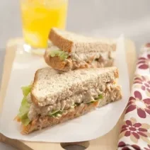 Foto em tons claros da receita de sanduíche de atum com queijo cottage servida em duas metades, uma por cima da outra, sobre uma tábua de madeira coberta com papel branco