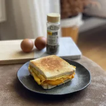 Foto de um sanduíche com ovo, sobre um prato preto em uma mesa. Ao fundo há uma tábua de madeira com um pote de tempero e dois ovos marrons.