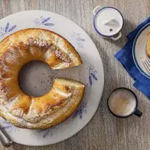 Fotografia em tons de cinza, branco e azul de uma bancada cinza e um prato banco com detalhes azuis, sobre ele o bolo de milho. Ao lado duas xícaras de café com leite e um paninho azul com um prato branco de bordas azuis com uma fatia do bolo e um garfo.