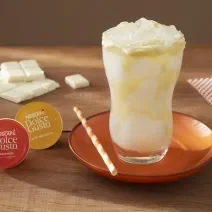 Foto da receita de mocha branco com latte macchiato servida em um copo de vidro alto sobre um pratinha laranja em cima de uma mesa de madeira com uma barra de galak e um paninho ao fundo