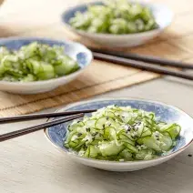 Fotografia em tons de verde em uma bancada de madeira com uma toalha de palha bege. Ao centro, um prato japonês branco com detalhes em azul e a salada de sunomono (pepinos fatiados bem finos) dentro, com um par de hashi apoiado.