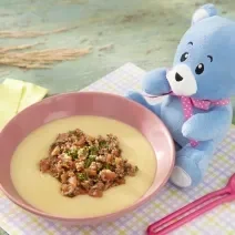 Foto da receita de Polenta Mole com Molho de Carne Moída. Observa-se um potinho rosa bebê com a polenta e a carne dentro, sobre um paninho quadriculado colorido. O ursinho Bo está do lado direito.