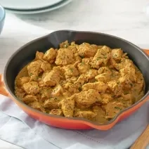 Foto da receita de frango ao curry sem lactose servida em uma frigideira grande laranja sobre uma mesa clara com um pano azul embaixo e uma colher de madeira ao lado