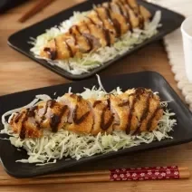 Foto da receita de Tonkatsu (Bife de Porco Japonês). Observa-se dois pratos retangulares pretos com repolho fatiado e o tonkatsu por cima, servido com um pouco de molho escuto. Observa-se um hashi na foto, na frente do prato.