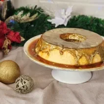 Fotografia em tons de dourado em uma bancada com um pano dourado e enfeites de natal. Ao centro uma bandeja redonda com um pudim de leite Moça e uma calda de caramelo com corante dourado comestível em cima.