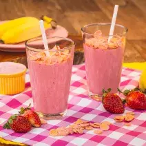 Imagem de uma bancada de madeira com um pano quadriculado em tom de rosa claro, morangos, e cereais espalhados, além de dois copos com uma bebida cor de rosa e canudos.