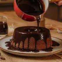 Fotografia em tons de vermelho em uma mesa de madeira com um prato branco com o bolo de chocolate em cima e uma calda de chocolate por cima. Uma panela vermelha em cima do bolo derramando a calda de chocolate.