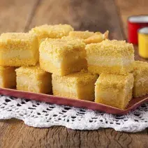 8 pedaços quadrados de bolo amarelo com uma lista branca no meio, dispostos em um prato vermelho que está em cima de uma toalha branca