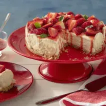 Fotografia em tons de vermelho em uma bancada de madeira de cor branca. Ao centro, uma boleira vermelha contendo a torta holandesa. Ao lado, há um pires com uma fatia da torta e um pano listrado vermelho.