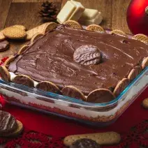 Foto da receita de Torta Holandesa na Travessa, em uma bancada de madeira decorada com temas de natal, alguns biscoitos espalhados, pedaços de chocolate branco. A receita tem um creme marrom sobre um creme claro e está em um refratário de vidro