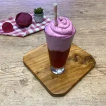 Fotografia em tons de rosa em uma bancada de madeira clara com uma tábua de madeira ao centro e um copo de vidro alto com uma bebida rosa e uma espuma rosa. Ao fundo, um pano xadrez rosa com uma pitaya aberta na metade e um limão-siciliano.