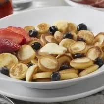 Fotografia mostra mini panquecas servidas em um prato branco, acompanhadas de morango e mirtilo