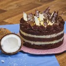 Foto da receita de Bolo Naked de Prestígio, decorado com pedaços de chocolate, sobre um prato rosa, numa bancada de madeira com tecidos azuis e dois pedaços de coco fresco aberto