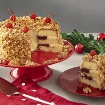 Foto com decoração natalina da receita de bolo de coroa de frankfurt servida sobre uma bailarina vermelha com um pedaço cortado ao lado sobre um prato de porcelana vermelho. Tudo isso em cima de uma mesa branca