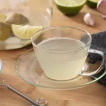 Fotografia em tons de verde em uma bancada de madeira, um pano cinza escuro, um prato de vidro pequeno com uma xícara e o chá de limão com alho dentro dele. Ao fundo, limão e alho espalhado pela mesa.