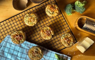 Fotografia vista de cima de uma mesa amarela com pedaços de brócolis cru, ralador, um pedaço de queijo e algumas forminhas de tom marrom. Sobre a mesa tem também uma grade preta e um pano azul, apoiado na grande tem seis muffins integrais de brócolis Fotografia vista de cima de uma mesa amarela com pedaços de brócolis cru, ralador, um pedaço de queijo e algumas forminhas de tom marrom. Sobre a mesa tem também uma grade preta e um pano azul, apoiado na grande tem seis muffins integrais de brócolis