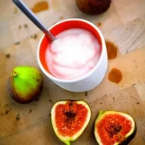 Fotografia de um creme de figo com creme de leite em um recipiente de cerâmica na cor branco e vermelho. Ao redor, dois figos inteiros e mais duas metades do fruto ao lado.