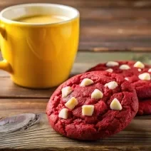 Fotografia de três cookies de red velvet com gotas de chocolate branco juntos, sobre uma mesa de madeira escura, ao lado de uma xícara grande amarela.