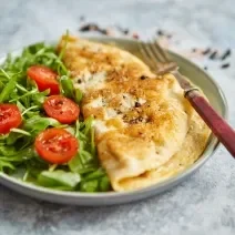 Foto da Receita de Omelete Baveuse. Observa-se um prato com a omelete dobrada no meio, servida com salada de rúcula e tomatinhos cerejas cortados ao meio.