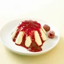 Fotografia de um pudim tradicional com calda de geleia de morango sobre um prato branco, pequeno e raso, e duas framboesas ao lado. O flan está sobre uma toalha de mesa na cor amarelo, em tom pastel.