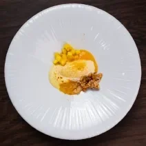 Fotografia vista de cima de um prato grande raso e branco com sorvete de tapioca com lírio-do-brejo, manga, curry e crocante de castanha com rapadura. O prato está sobre uma bancada marrom de tom escuro.