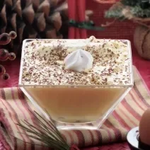 Fotografia de um flan de NESCAFÉ ao molho zabaione dentro de um recipiente quadrado de vidro fundo, com raspas de chocolate e chantilly por cima. A sobremesa está sobre um pano de mesa colorido.