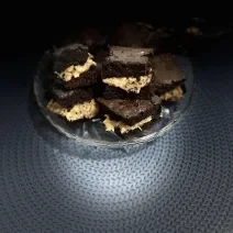 Fotografia em tons escuros de pedaços de brownies recheado, um sobre o outro, e apoiados em um apoio de vidro, sobre um pano de lã azul.