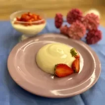 Fotografia em tons de rosa e azul com um prato rosa ao centro. Em cima do prato existe uma receita de Panna Cotta com morangos fatiados por cima