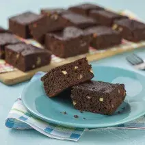 fotografia em tons de azul e marrom de uma bancada vista de frente, contém um pano em tons de branco e azul, um prato azul e redondo contém 2 Brownies e ao fundo um tábua de madeira com diversos Brownies