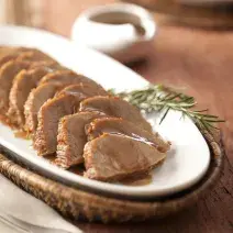 foto em tons marrom e branco. Uma mesa de madeira contém um cesto com um prato que comporta pedaços de carne fatiados.