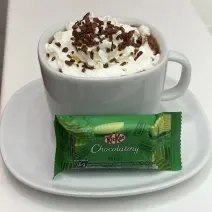 Fotografia de uma xícara branca com um hot chocolate com chantilly e granulado por cima. Na frente da xícara tem um kitkat de menta, e está sobre um prato pequeno de vidro raso branco.