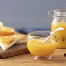 Fotografia em tons de laranja em uma bancada de madeira, uma tábua de madeira, um paninho azul, um pote hermético de vidro com geleia de laranja dentro. Ao lado, um potinho de vidro com mais geleia. Ao lado, fatias de pão e laranjas cortadas ao meio.