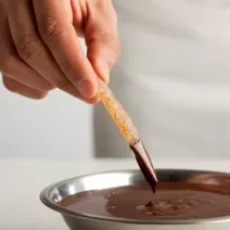 Fotografia de fruta seca afundando no chocolate amargo, que está em um recipiente de alumínio fundo. O qual está sobre uma mesa branca.