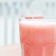 Fotografia em tons claros de uma bebida com um pouco de espuma de melancia com leite MOÇA e neston, que está em um copo de vidro.