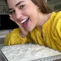 Fotografia em tons de amarelo em uma bancada com a travessa de torta de Farinha Láctea e a atriz e apresentadora Vivian Amorim aparecendo de fundo.