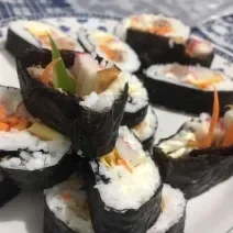 Fotografia de fatias grossas de gimbap brasileiro com arroz, cenoura, kani e pepino sobre um prato branco.