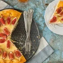 Fotografia vista de cima de uma torta de amêndoas com creme de leite e figos, coberta com figos fatiados, sobre o fundo falso da forma e uma fatia ao lado