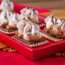 Fotografia em tons de vermelho com vários biscoitos Choco Biscuit ao centro. Por cima de cada biscoito existe uma camada de doce de leite, banana e, por cima, chantilly.