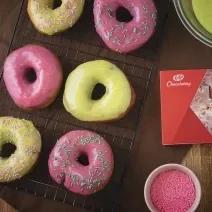 Foto da receita de Donuts Wicked, vistos de cima, servidos em uma grade de ferro preta. São seis no total, decorados com cobertura rosa e verde e alguns com granulados coloridos. Ao lado direito há uma caixa de KitKat Wicked.