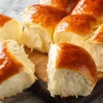 Fotografia de várias unidades pãos fofinhos de leite ninho dourados por cima. Os pães estão sobre uma tábua de madeira.