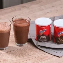 Foto da receita de smoothie pós treino de nescau extra cacau servida em dois copos de vidro altos sobre uma mesa de madeira com duas embalagens de nescau extra cacau 30% e 60% sobre um paninho cinza