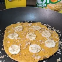 Fototgrafia de uma omelete de neston com rodelas de banana e neston cereias polvilhado, sobre uma frigideira.