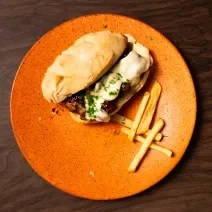 Fotografia vista de cima de um sanduíche de costelinha de porco com barbecue de frutas vermelhas e queijo, acompanhado de batatas fritas que está no mesmo prato, cujo a cor é laranja. O prato está sobre uma bancada de madeira.