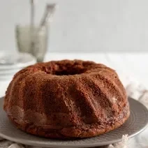 Fotografia de um bolo de chocolate feito em forma com furo central sobre um prato raso de vidro cinza. Ao lado tem granulado de chocolate em um prato pequeno raso. No fundo tem pratos brancos e garfo, e o bolo está sobre um pano branco.