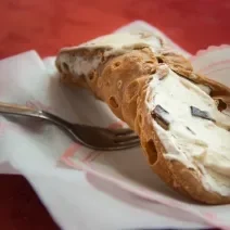 Fotografia de um cannoli de chocolate com creme branco sobre um papel, e ao lado de um garfo.