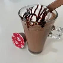 Fotografia em tons de branco com uma caneca de vidro ao centro. Dentro da caneca existe uma receita de frappuccino de cor chocolate coberto com chantilly e calda de chocolate. Ao lado existe uma cápsula de NESTLÉ Dolce Gusto KitKat