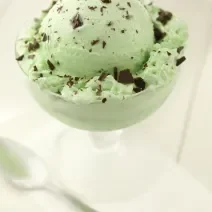 Fotografia de uma bola de sorvete de menta com chocolate em uma taça, que está sobre uma mesa branca. E, ao lado da taça, uma colher de sobremesa.
