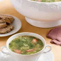 Foto de um recipiente redondo, como uma xícara, só que maior, com caldo verde dentro, em cima de um prato branco