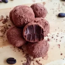 Fotografia de várias trufas de cacau e café passadas no cacau em pó. A trufa da frente foi mordida, e as trufas estão sobre uma tábua de madeira clara.